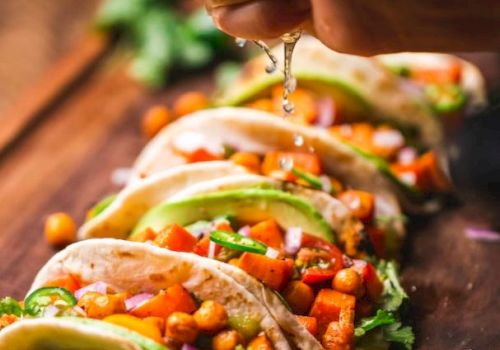 The image shows a hand squeezing lime juice over a row of tacos filled with chickpeas, diced tomatoes, onions, and garnished with cilantro and avocado.