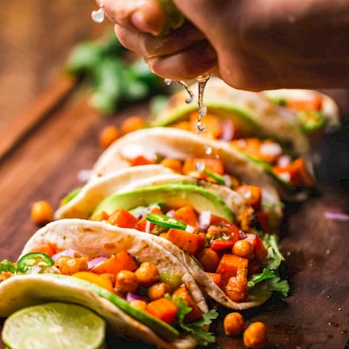 The image shows a hand squeezing lime juice over a row of tacos filled with chickpeas, diced tomatoes, onions, and garnished with cilantro and avocado.