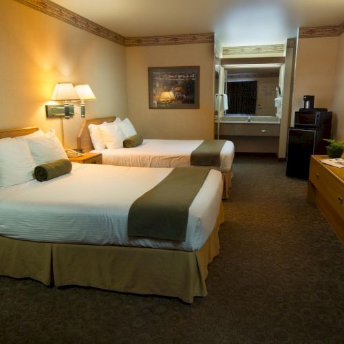 The image shows a hotel room with two neatly made beds, lamps, a TV, a dresser, and a small kitchenette area with a microwave and mini fridge.
