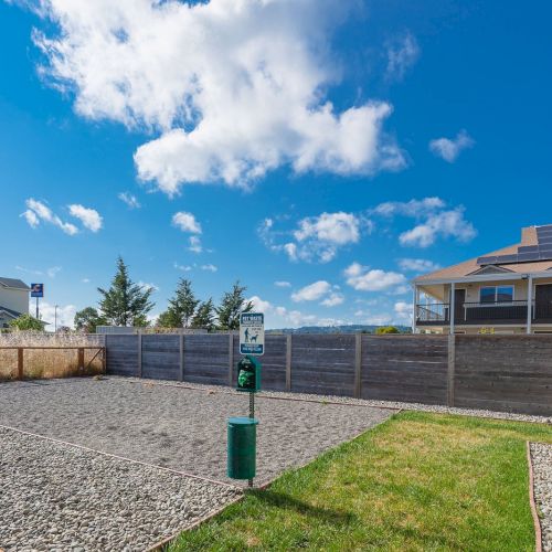 A fenced area with a gravel surface, pet waste station, and buildings in the background. Blue sky with clouds above.