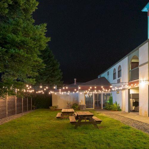 A nighttime courtyard scene with picnic tables on grass, string lights overhead, and a building with arched windows and entrances.