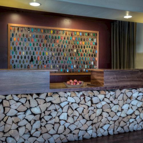 A reception desk with stacked wood in front and a colorful key holder board behind, flanked by lamps and pomegranates on the counter.