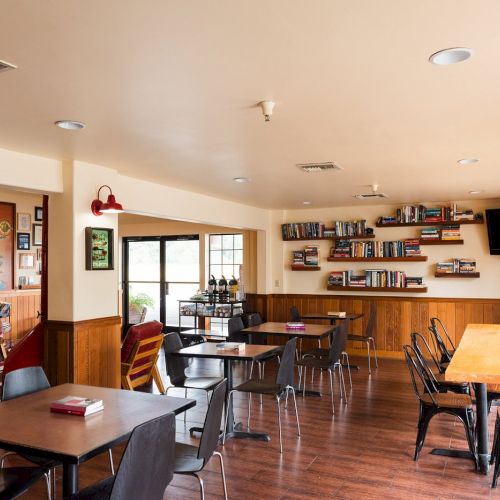 A cozy library café with wooden floors, bookshelves, tables, chairs, and wall art, creating a relaxed atmosphere.