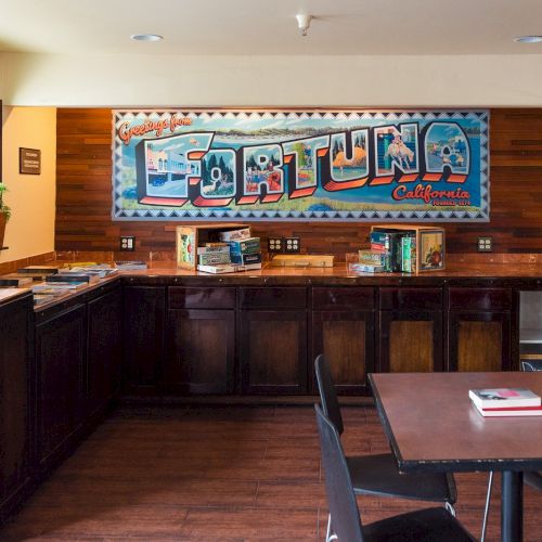 A room with a "Greetings from Fortuna, California" sign, board games on a counter, a plant, and water with lemons.
