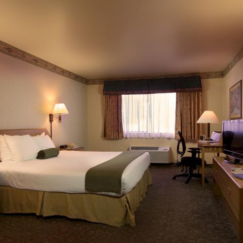A cozy hotel room featuring a neatly made bed, two bedside lamps, a TV, desk with chair, window, artwork, and wall decorations.