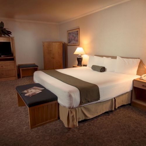 A cozy hotel room with a large bed, two lamps, a TV, wardrobe, and a bench. Neutral colors create a warm and inviting ambiance.