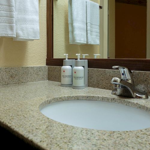 A bathroom countertop with a sink, faucet, two white bottles labeled "The Redwood", and white towels on a rack above.