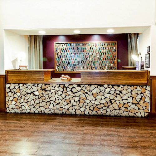 A reception desk with a wooden log facade, modern lighting, and a colorful mosaic artwork in the background, on a wooden floor.