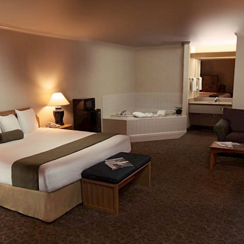 The image shows a cozy hotel room with a king-size bed, a small sofa, a desk with a laptop, and a hot tub near the bathroom area.