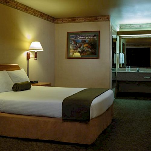 A hotel room with a neatly made bed, bedside tables with lamps, a painting, a wardrobe, and a vanity area with a mirror in the background.