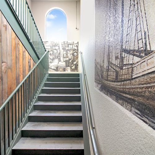 A stairway with metal railings, a wood-paneled wall, a maritime-themed mural, and a window at the top showing a blue sky and clouds.