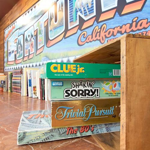 The image shows a stack of board games, including CLUE Jr., Sorry!, Trivial Pursuit, and The 80’s Game, against a wall with 