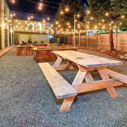 The image shows an outdoor area with wooden picnic tables, string lights overhead, and a gravel ground. Trees and a fence line the area.