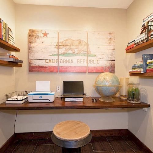 A small home office with a desk, laptop, printer, globe, and bookshelves. The wall features a large California Republic flag art piece.