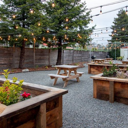 An outdoor patio with picnic tables, wooden planters, string lights, and trees in the background. The ground is covered with gravel, creating a cozy atmosphere.