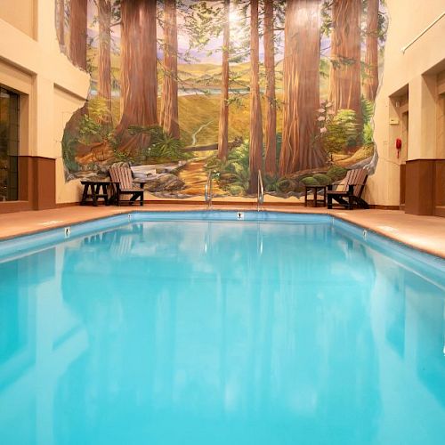 An indoor swimming pool with clear blue water surrounded by lounge chairs, trees mural, and doors on the sides.