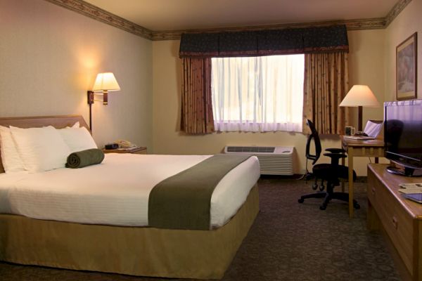 A cozy hotel room with a large bed, TV, desk, chair, bedside lamps, window, and decor, creating a welcoming atmosphere for guests.