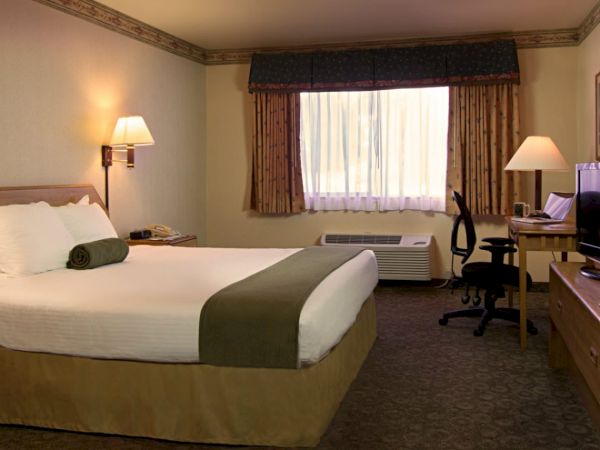 This image shows a well-lit hotel room with a large bed, bedside tables, lamps, a desk, a TV, and a window with curtains ending the sentence.