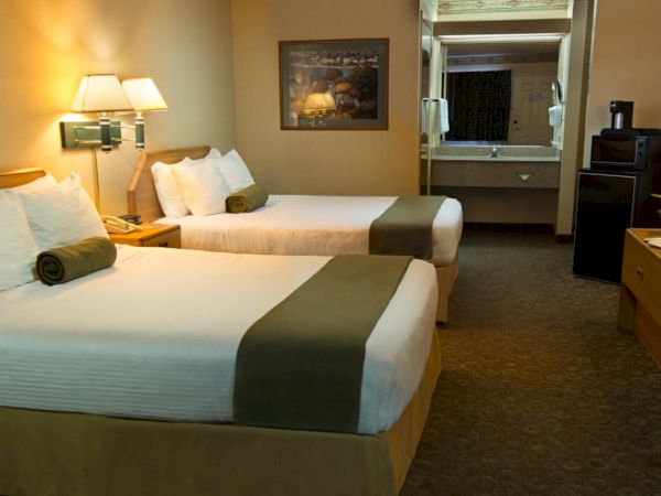 This is a hotel room with two neatly made beds, bedside lamps, a TV, and a small fridge. A bathroom or vanity area can be seen in the background.
