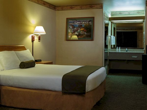 A cozy hotel room with a neatly made bed, lamps, painting, nightstand with a phone and an open space leading to a sink area.