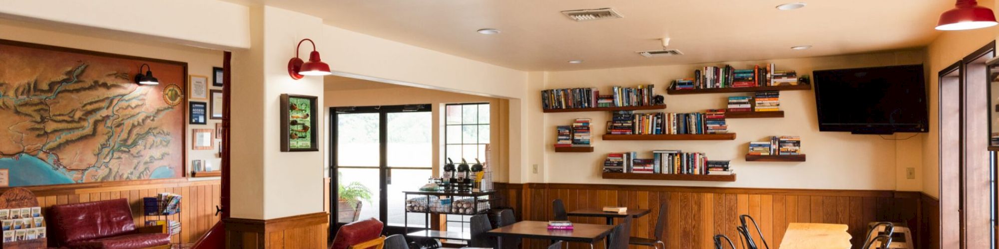 The image shows a cozy cafe or restaurant with tables, chairs, wall-mounted bookshelves filled with books, a TV, and modern decor.