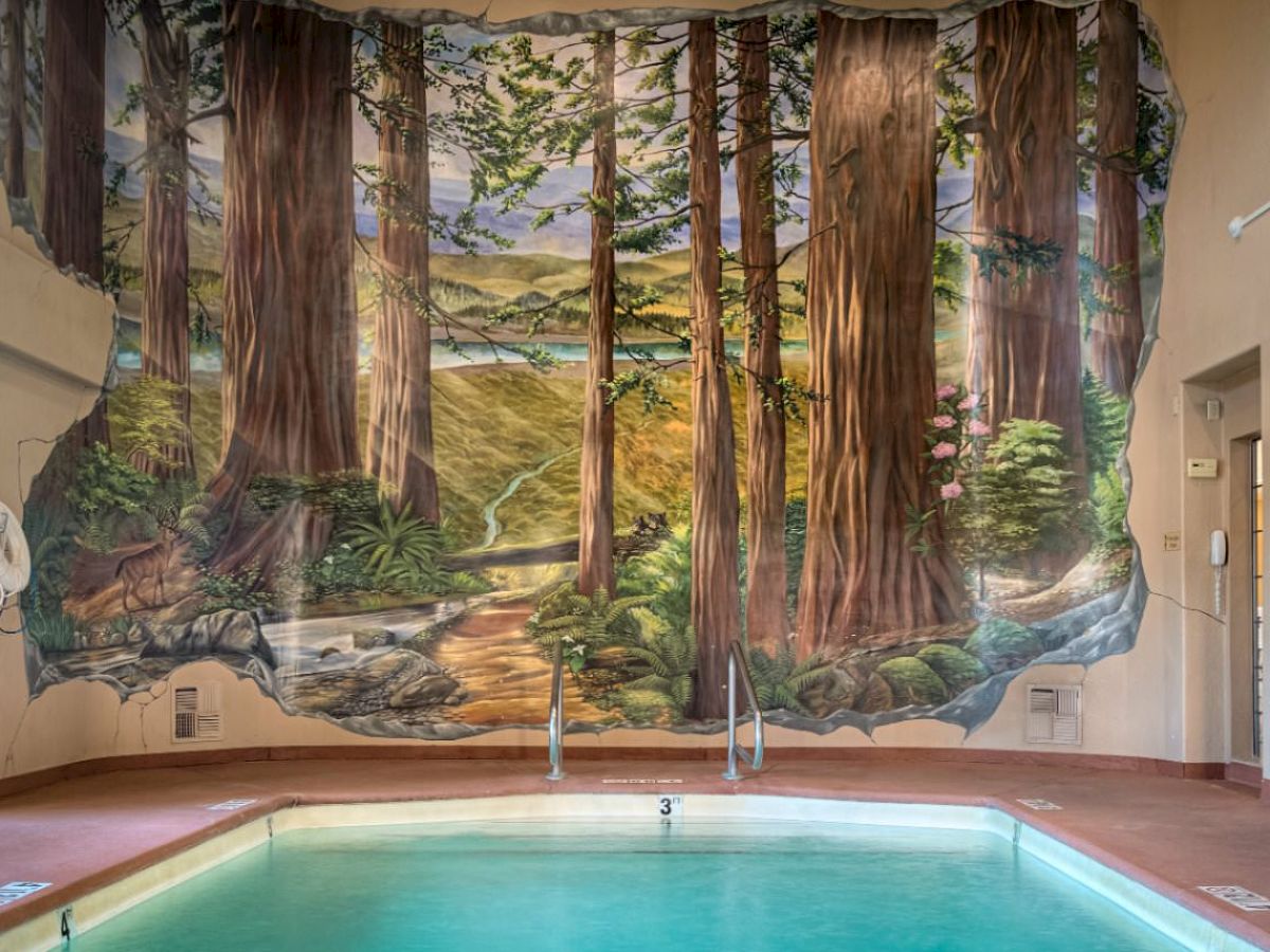 An indoor pool with a colorful mural depicting a forest and a pathway. Large windows allow natural light to enter the room.