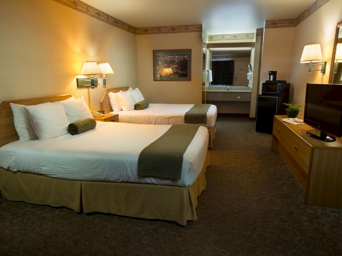 The image shows a hotel room with two double beds, nightstands, lamps, a TV on a dresser, a mirror, and a small kitchenette area with a sink.