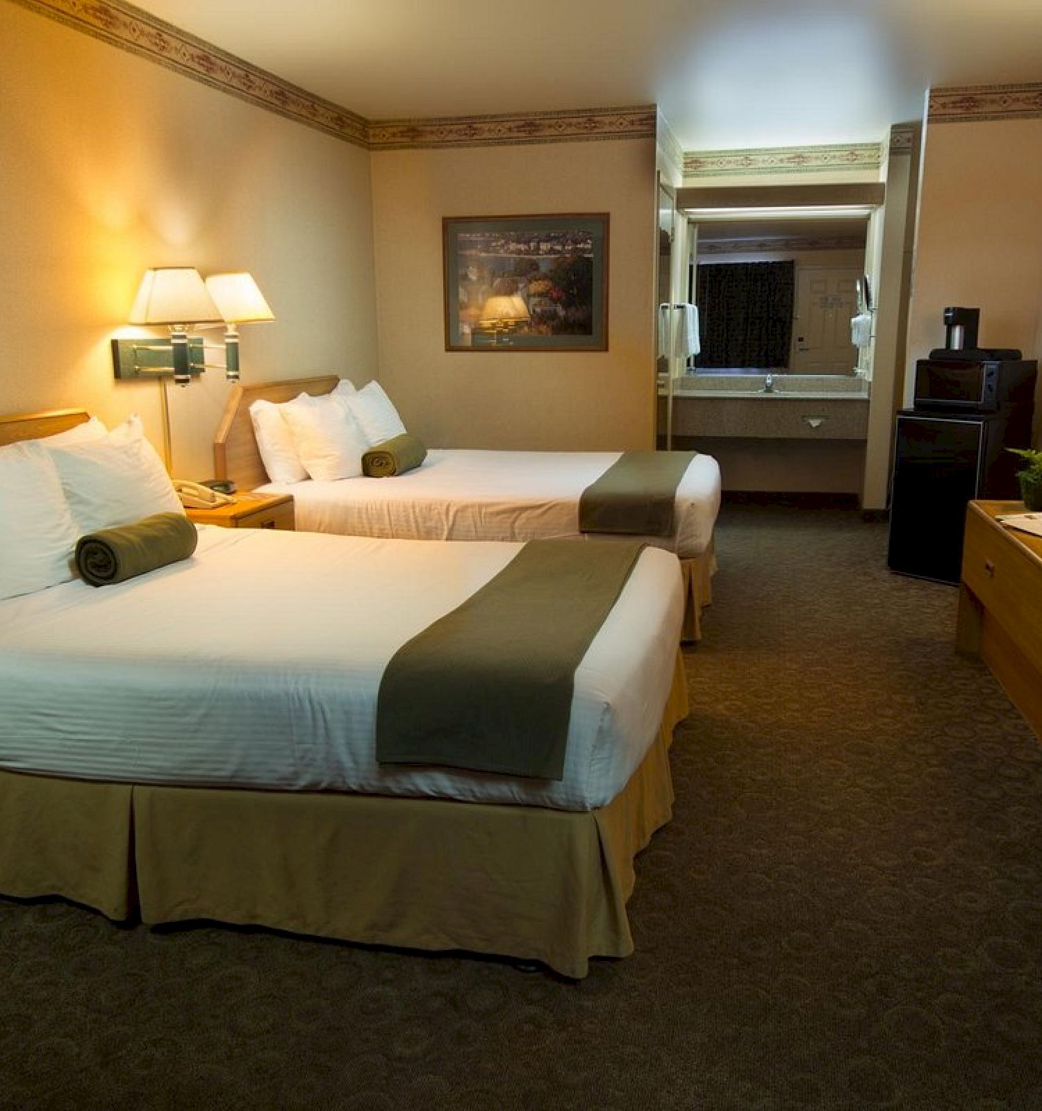 The image shows a hotel room with two double beds, nightstands, lamps, a TV on a dresser, a mirror, and a small kitchenette area with a sink.