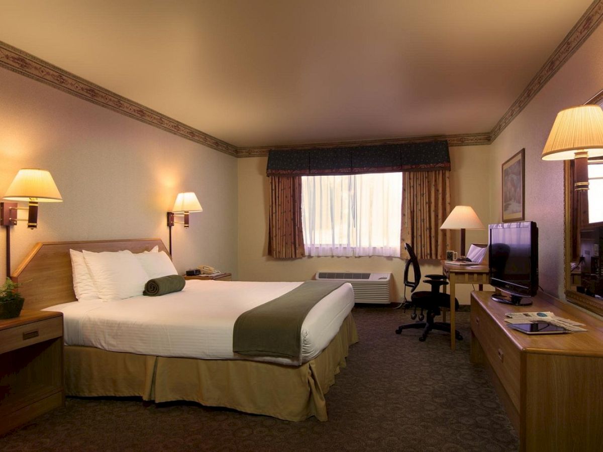 A hotel room with a double bed, bedside tables, lamps, a desk, a chair, a TV, and a window with curtains.