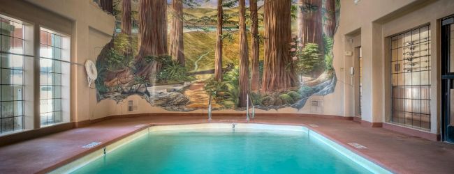 An indoor pool with blue water and a mural of a forest scene on the far wall, surrounded by windows and doors.