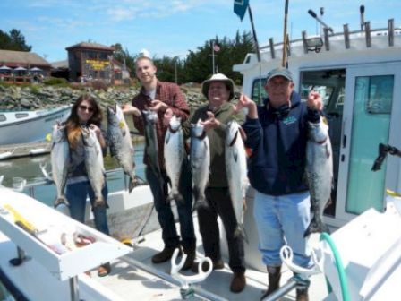 Four people on a boat proudly holding up several large fish each, with a coastal background and buildings visible.