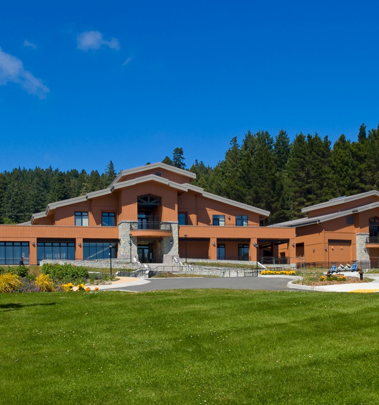 A modern building with multiple rooflines, surrounded by landscaped greenery, trees in the background, and a bright blue sky.