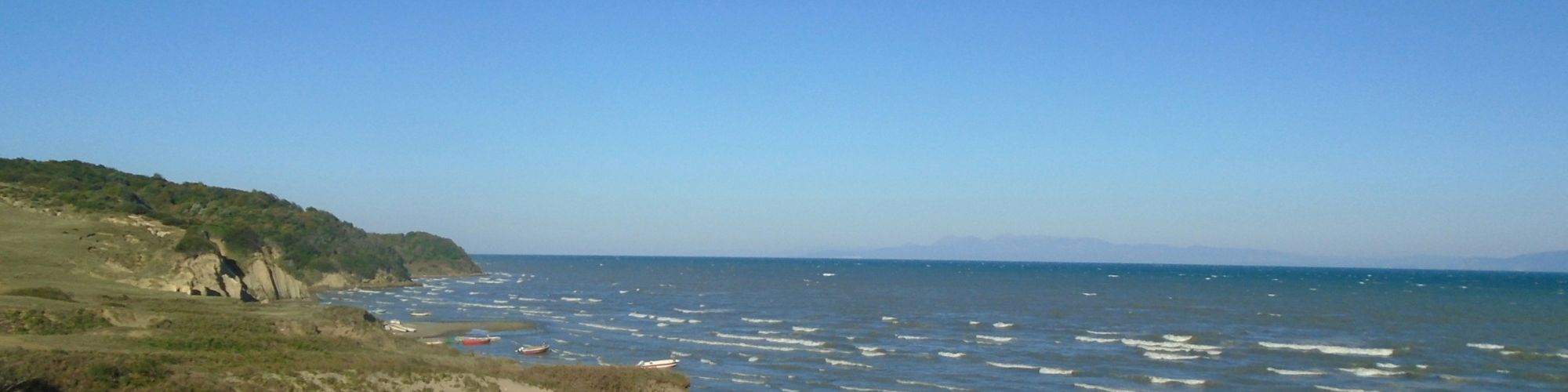 A coastal landscape with a sandy beach, scattered waves, and distant hills under a clear blue sky.