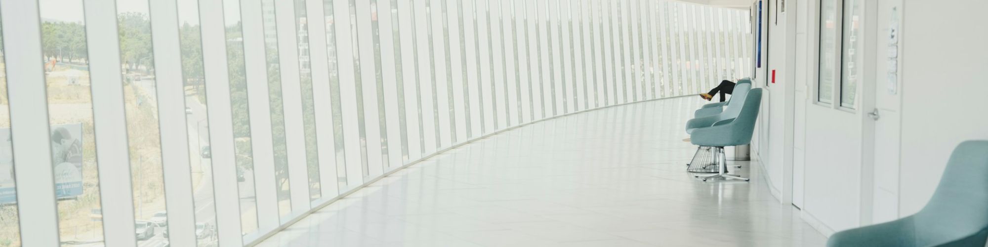 A bright, modern hallway with large windows and blue chairs along the wall, creating an airy, spacious atmosphere.