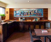 A retro diner counter with wood panels, a bright mural behind the counter, a few stools, a window, plants, and a small table set in front.