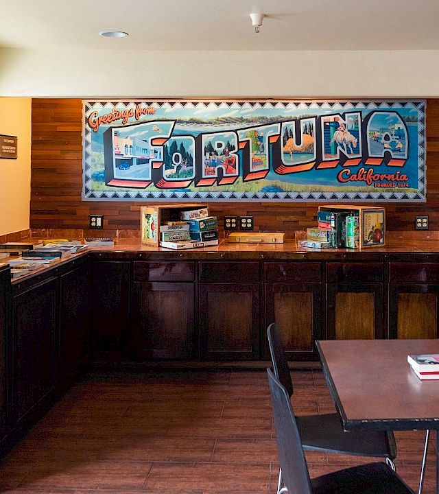 A retro diner counter with wood panels, a bright mural behind the counter, a few stools, a window, plants, and a small table set in front.