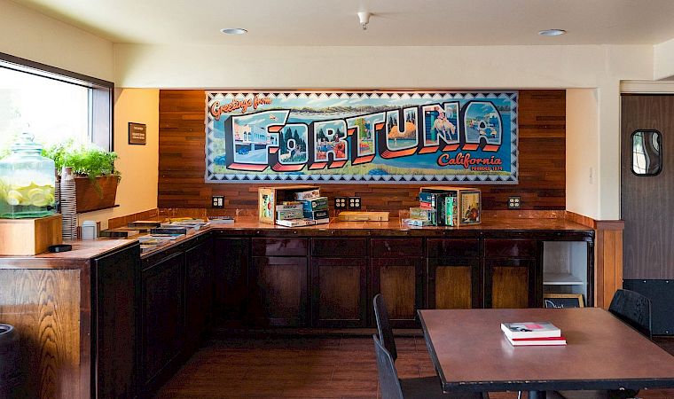 A retro diner counter with wood panels, a bright mural behind the counter, a few stools, a window, plants, and a small table set in front.