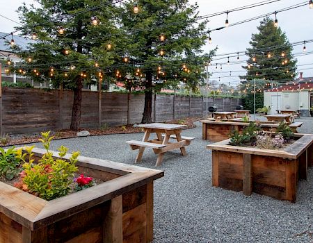 An outdoor rustic seating area with wooden planters, picnic tables, string lights, gravel ground, and trees in a cozy yard.