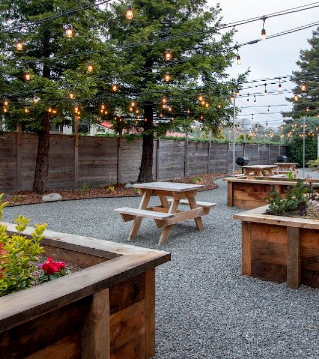 An outdoor rustic seating area with wooden planters, picnic tables, string lights, gravel ground, and trees in a cozy yard.